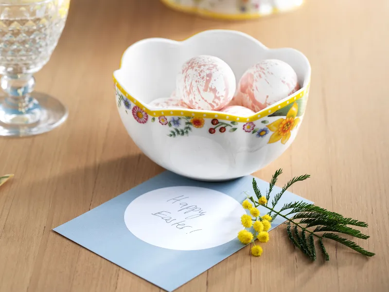 Un plat creux décoratif Spring Awakening avec des œufs de Pâques peints sur une table en bois, accompagné d’une carte de vœux portant l’inscription « Frohe Ostern ! » (c’est-à-dire « Joyeuses Pâques ») et d’une branche de fleurs jaunes.