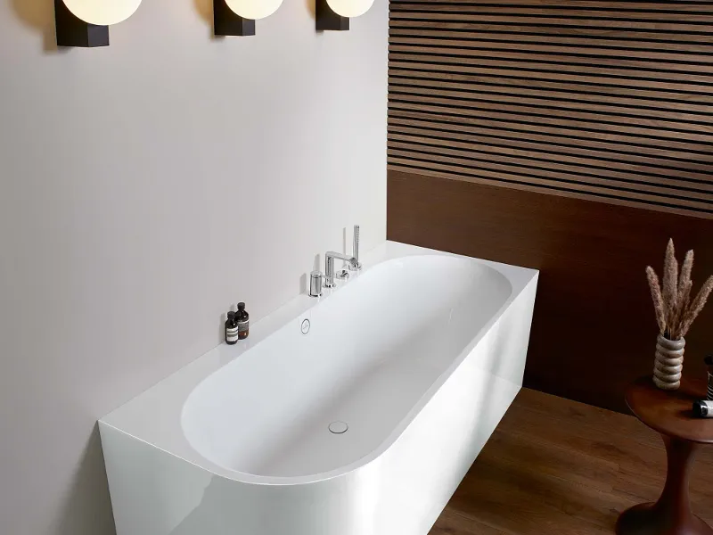 Bagno moderno con vasca free-standing bianca e verde, tre applique sferiche sovrastanti e accenti in legno su pareti e pavimento. Un vaso con rami decorativi si trova su un tavolino.