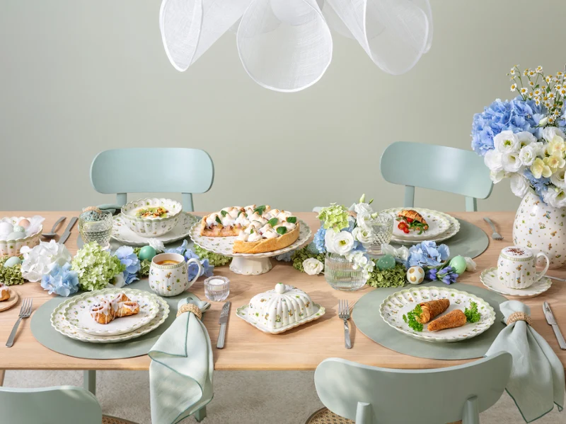 Une table à manger pour quatre personnes décorée dans des tons pastel, avec des assiettes remplies de plats, des tasses à thé, des compositions florales bleues et blanches et une vaisselle élégante, créant une atmosphère fraîche et accueillante.