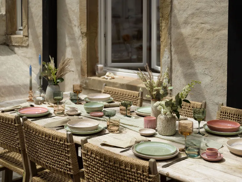 Tavolo da pranzo rustico per esterni con sedie intrecciate, piatti Perlemor color pastello di like. by Villeroy & Boch, bicchieri e centrotavola floreali sullo sfondo di un muro di pietra con finestra.