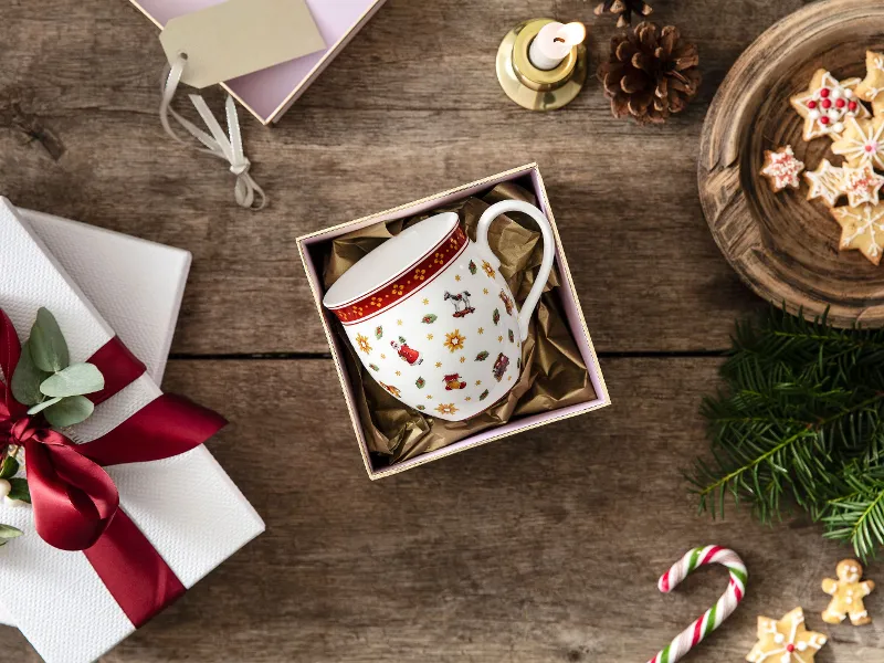 Una tazza Toy's Delight in una scatola regalo, circondata da un regalo incartato bianco con un fiocco rosso, biscotti su un piatto di legno.