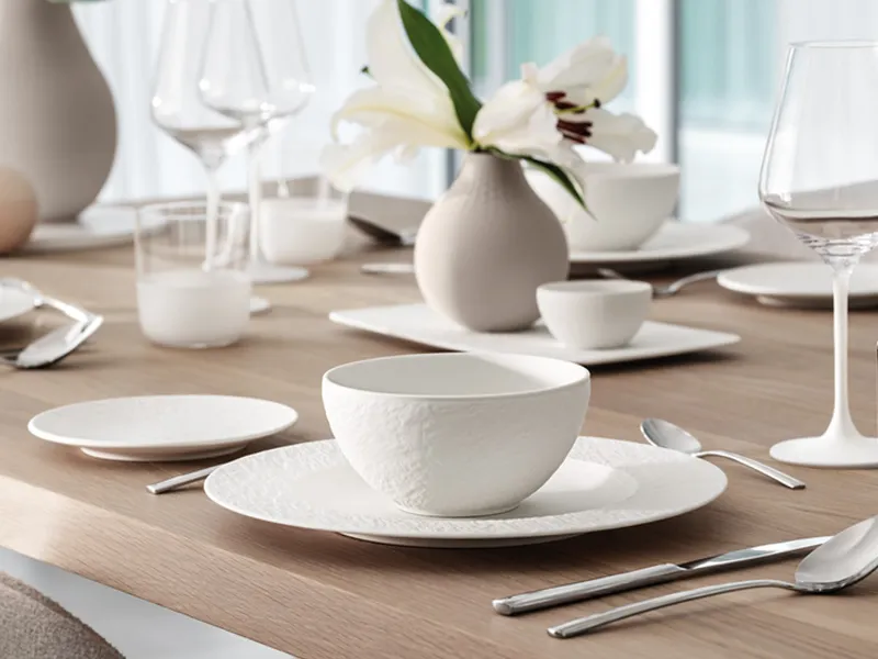 Elegant table decoration with white crockery, glasses and cutlery on a wooden table. A vase with white flowers serves as a centrepiece.