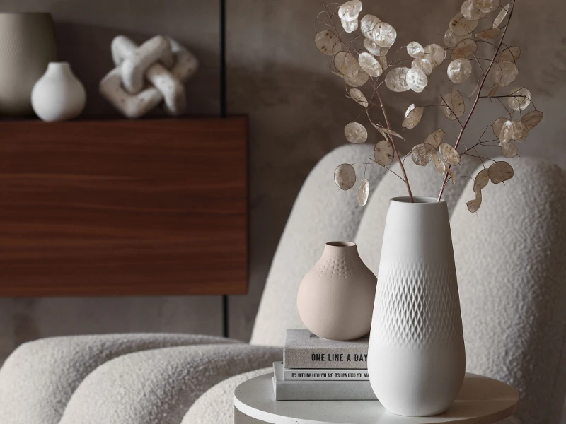 Une table moderne avec un vase Manufacture blanc contenant des fleurs séchées, des livres et des objets décoratifs à côté d’une chaise moelleuse dans des tons neutres, dans un intérieur moderne.