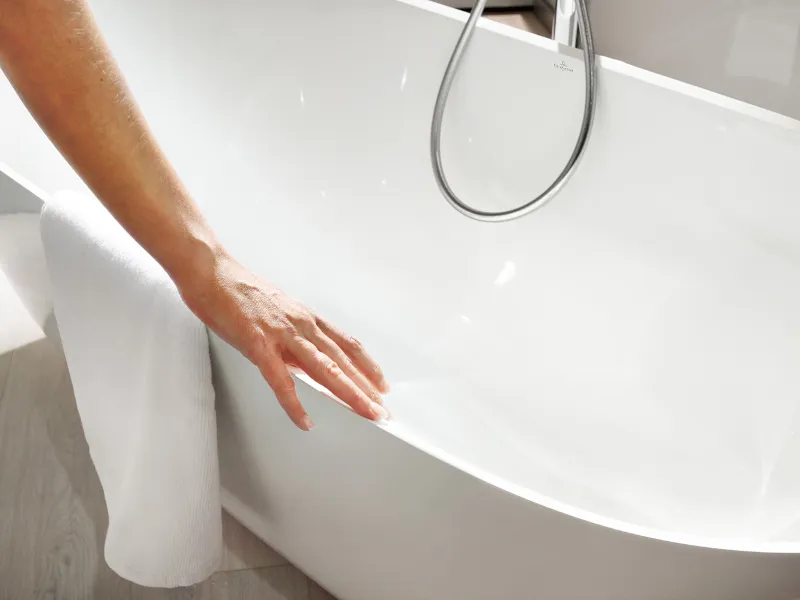 Die Hand einer Person berührt den Rand einer weißen Badewanne, an deren Seite ein Handtuch hängt.