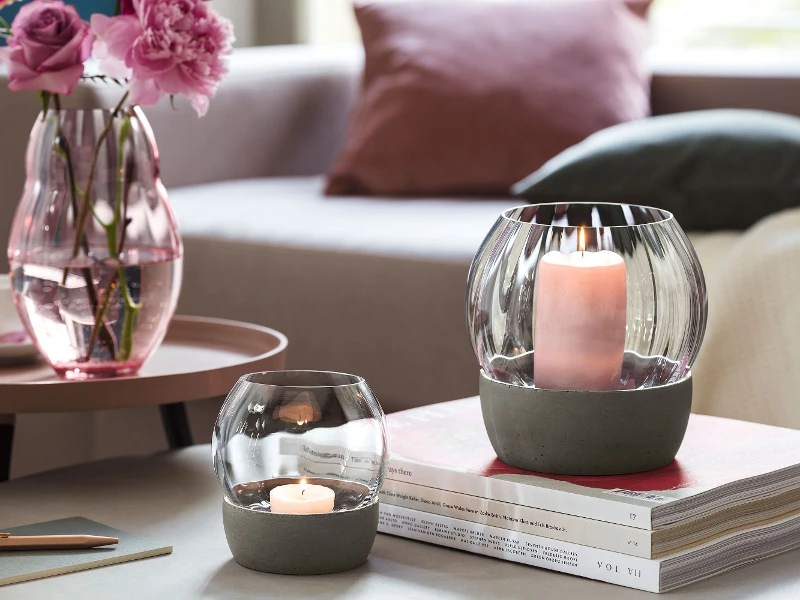 Un salon confortable avec un canapé rose, un vase en verre Rose Garden rempli de fleurs, des bougies allumées dans des supports en verre Rose Garden, du café et des livres sur une table ronde.