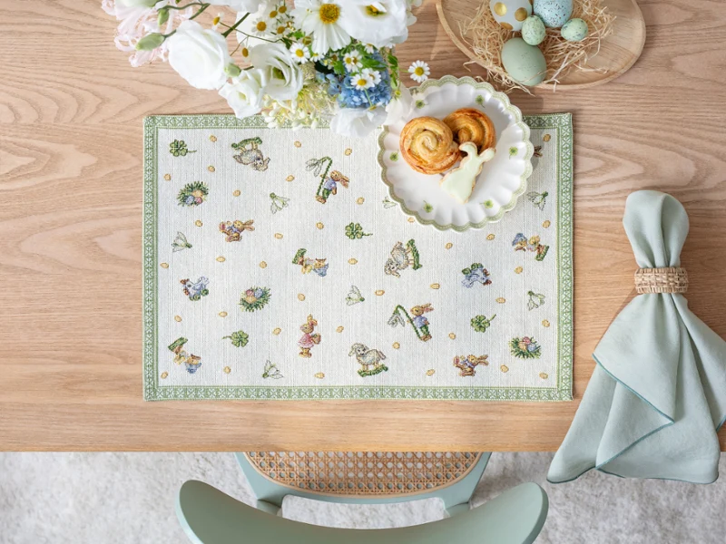 Une table en bois dressée avec un set de table Easter Delight, des pâtisseries sur une assiette, des fleurs, des œufs teints et une serviette pliée.