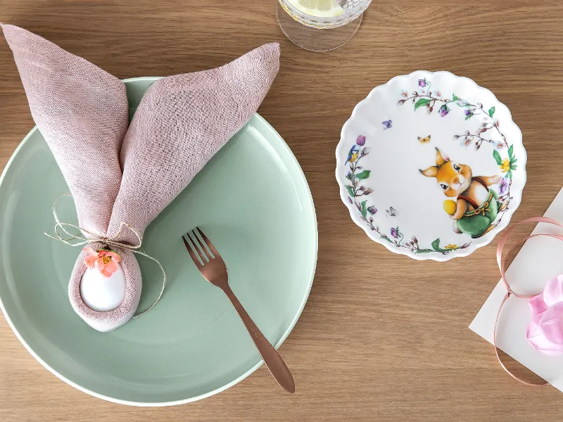 Ein grüner Teller „it‘s My Match“ von Villeroy & Boch dekoriert mit einer rosa Serviette, einem weißen Ei, einer braunen Gabel und einem dekorativen Spring Fantasy Blumenteller mit einem Häschenfigur auf einer Holzoberfläche.