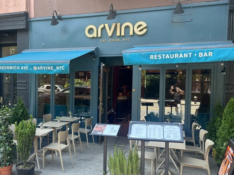 The storefront of Arvine restaurant in NYC, featuring a dark teal facade, blue awnings, outdoor cafe seating, and menu stands on the sidewalk.