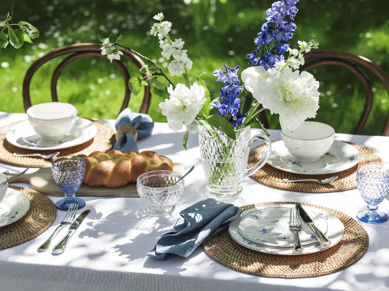 Une table de jardin est dressée avec de la porcelaine fleurie « Vieux Luxembourg » de Villeroy & Boch, des sets de table en osier, de la vaisselle en verre teinté bleu, un pot en cristallin avec des fleurs bleues et blanches et une miche de pain.