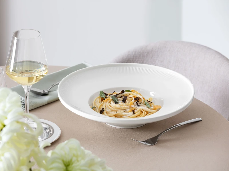 Afina pasta bowl from Villeroy & Boch with tagliatelle, sage and white sauce, next to a glass of white wine and Piemont flatware.