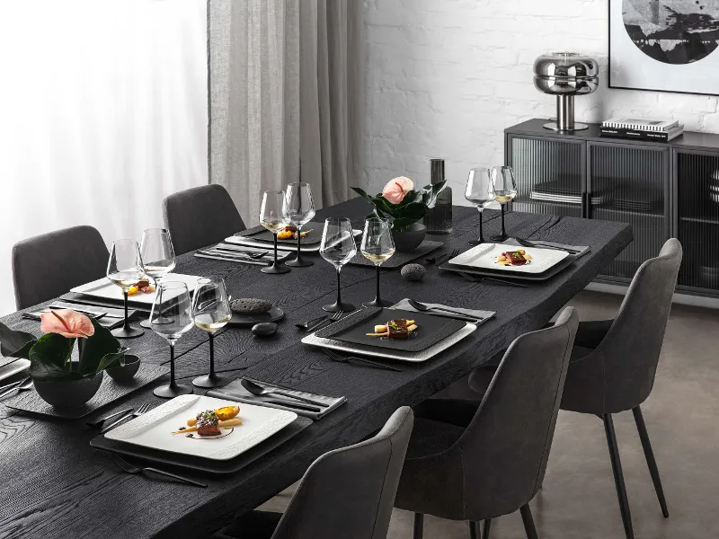 Ein schwarzer Tisch in einem eleganten Wohnzimmer gedeckt mit Gläser, rechteckigem Geschirr und Besteck aus der Kollektion Manufacture von Villeroy & Boch.