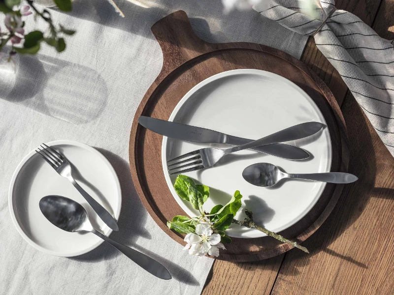 Elegant table decor with simple white plates and Arthur brushed stainless steel flatware, accentuated by natural light and a sprig of flowers.