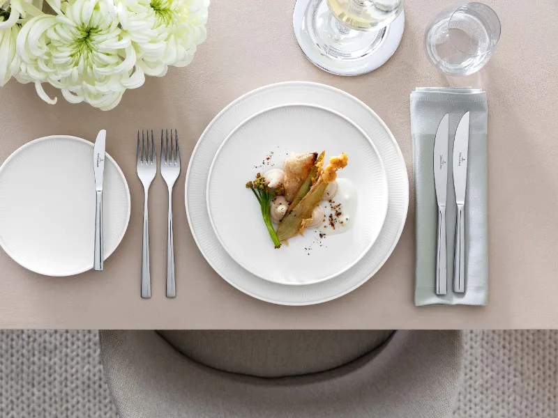 Une décoration de table élégante avec une assiette gourmet Afina blanche, des couverts de table Piemont et une décoration florale.