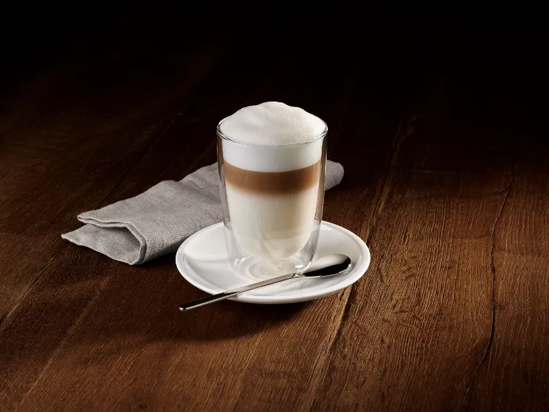 A glass of layered latte with foam on a Villeroy & Boch Coffee Passion saucer, together with a spoon and next to a folded grey napkin on a wooden surface.