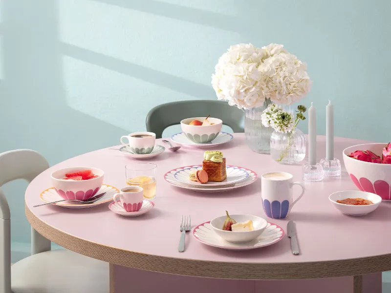 Un tavolo rotondo con piatti, tazze e ciotole piene di cibo, decorato con motivi rosa. Al centro c'è un vaso di fiori bianchi e due candele spente. Le sedie circondano il tavolo.	