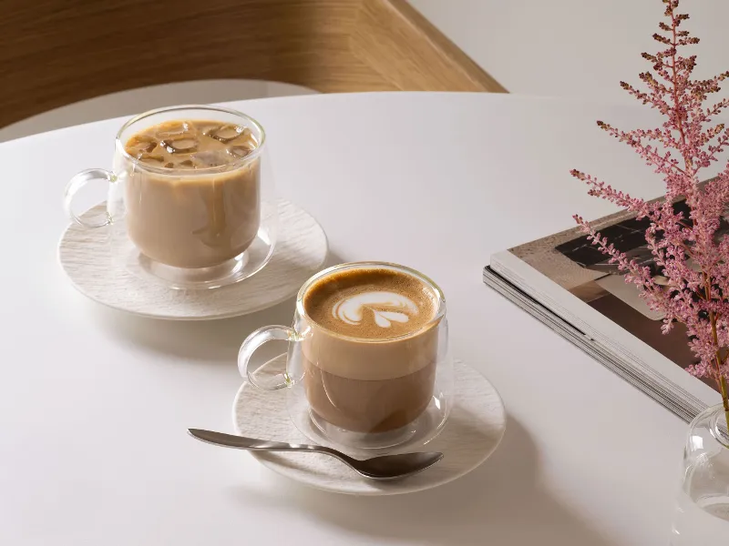 Deux tasses en verre Artesano de Villeroy & Boch sur une table : l’une remplie de café glacé, l’autre de latte, avec une cuillère, une pile de magazines et une fleur rose dans un vase.