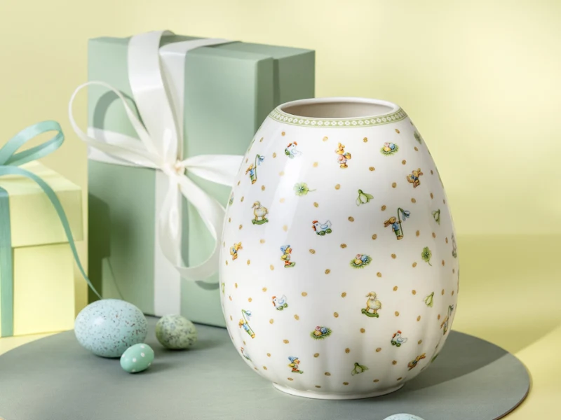 Un vase blanc Easter Delight avec des motifs floraux est exposé à côté de deux boîtes-cadeaux aux couleurs pastel et d'œufs mouchetés.
