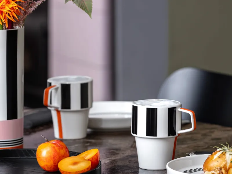Modern table setting with a striped Memphis vase, matching cups, fruit and a bread roll on a dark marbled background.