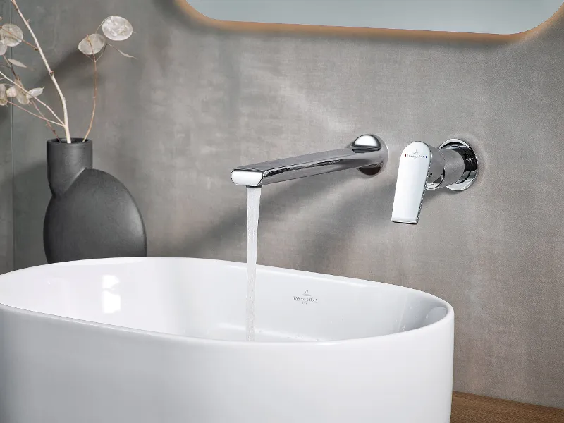 Un moderno lavabo nel bagno, con l'acqua che sgorga dal rubinetto cromato. Un vaso nero con piante secche si trova accanto al lavabo Collaro di Villeroy & Boch.