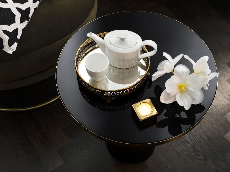 Une théière MetroChic blanche avec une tasse assortie, deux fleurs blanches et une bougie allumée sont posées sur une table ronde noire avec un coussin à motifs en arrière-plan.