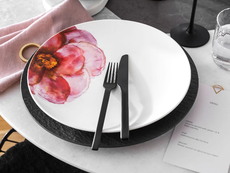 Table decoration with a Rose Garden plate with floral decoration on a black Manufacture plate and black flatware and a menu next to it.