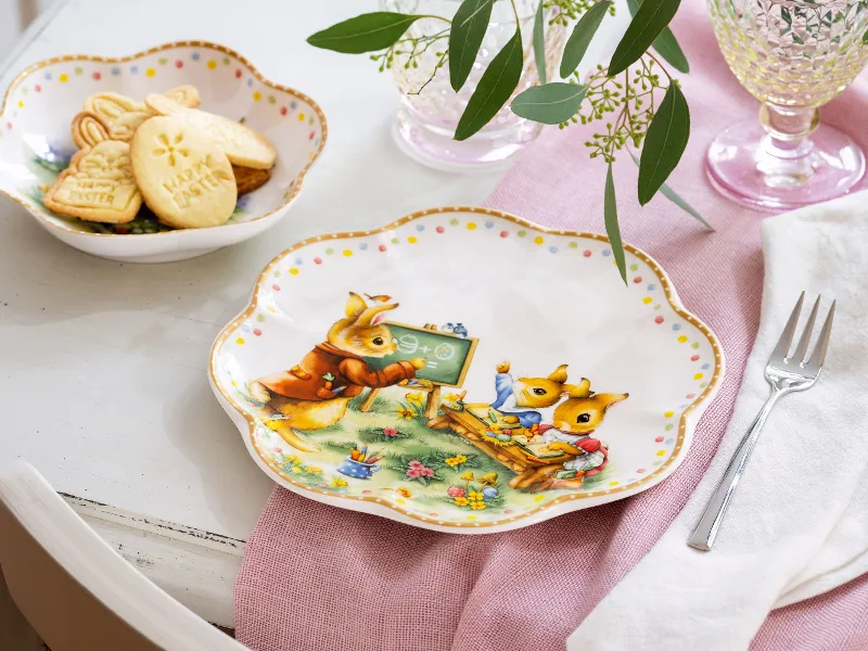 Die dekorativen Teller der Annual Easter Edition von Villeroy & Boch mit Häschen sind elegant auf einem Tisch mit einer rosa Serviette, einer Gabel und einer Schüssel Kekse arrangiert.