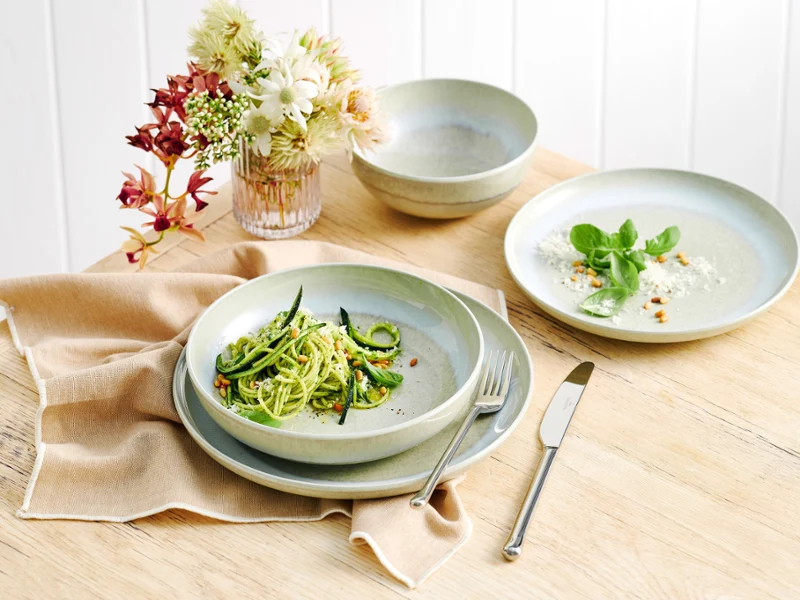 aus-perlemoralga-indoor-food-nopeople-dinnerset7069 The Perlemor Alga dinner set for one, featuring a plate filled with fresh zucchini pasta, rests on a brown linen tablecloth atop a rustic wooden table, accompanied with silver cutlery.