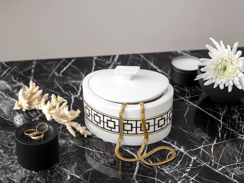 A decorative, gold-patterned MectroChic sugar bowl on a marble surface, surrounded by dried coral, a candle, a flower and a ring on a black lid.