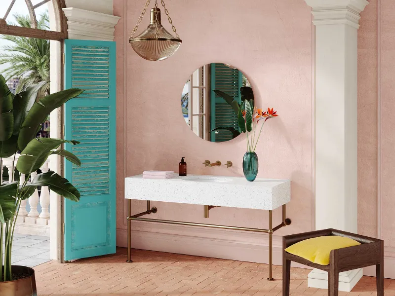 Bagno con pareti rosa, specchio rotondo, lavabo bianco Loop & Friends di Villeroy & Boch e rubinetteria in ottone. Persiane blu, pianta tropicale in vaso, sgabello in vimini con cuscino giallo e lampada a sospensione.