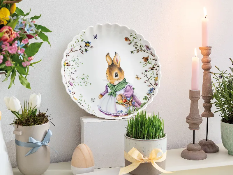 The Spring Fantasy decorative plate by Villeroy & Boch with a bunny motif is displayed on a shelf, surrounded by flowers, candles, and a wooden egg.