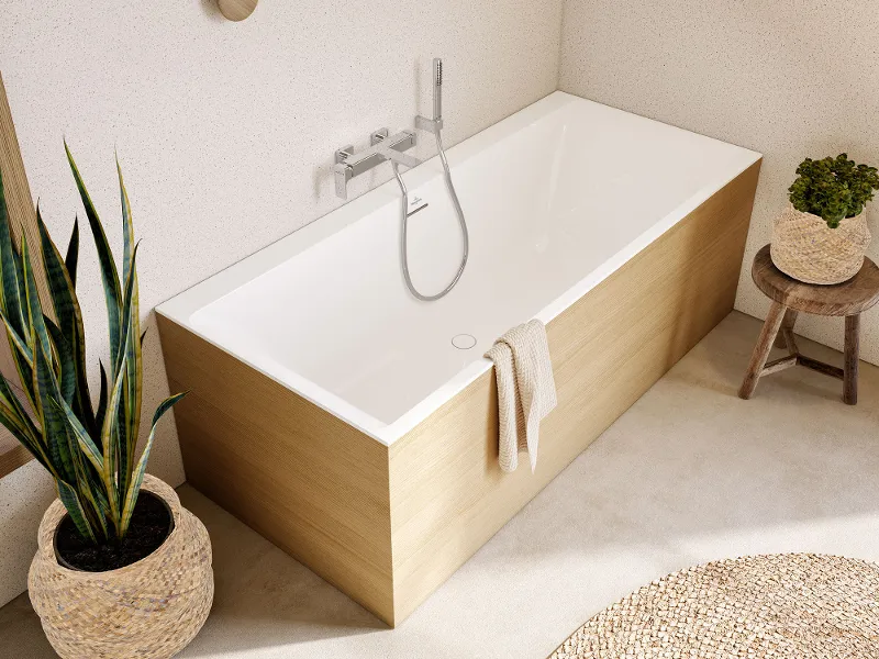 Een moderne badkamer met een rechthoekig bad uit de Subway collectie van Villeroy & Boch, een houten krukje met een plant, een geweven vloerkleed en een grote potplant naast het bad. De muren hebben een licht gevlekte afwerking met ronde houten accenten.