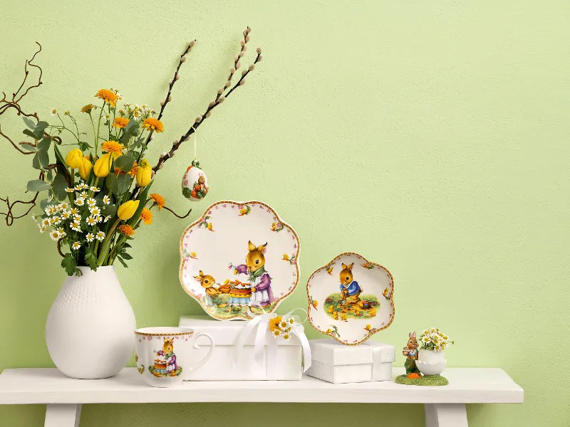 Sur un présentoir blanc se trouvent un vase avec des fleurs jaunes, deux assiettes décoratives, une tasse, une petite figurine et un ornement en forme d’œuf « Annual Easter Edition » de Villeroy & Boch. Le mur en arrière-plan est peint en vert clair.