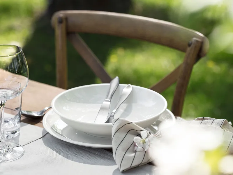 A table setting with a white Artesano plate, a bowl and cutlery, next to a wine glass and a napkin on a wooden table with a chair.