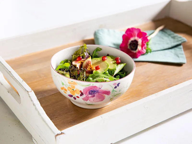 Una ciotola di insalata con verdure a foglia, semi di melograno e fichi su un vassoio bianco Mariefleur di Villeroy & Boch con motivo floreale. Sullo sfondo si vedono un fiore rosa e un tovagliolo verde.