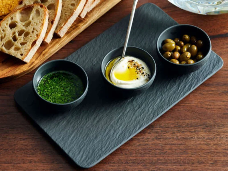 A slate platter with three small bowls contains green herbs, creamy dip with oil and pepper, and olives. Slices of bread are beside the platter.