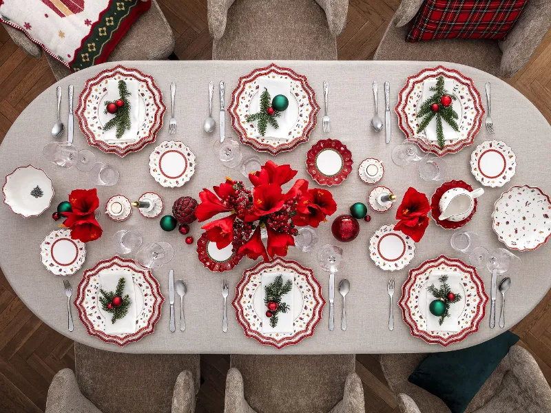 A festively decorated dining table for eight people with Villeroy & Boch Toy’s Delight red and white plates, arranged cutlery, red flowers, Christmas decorations and a grey tablecloth in a cosy dining room with grey chairs.