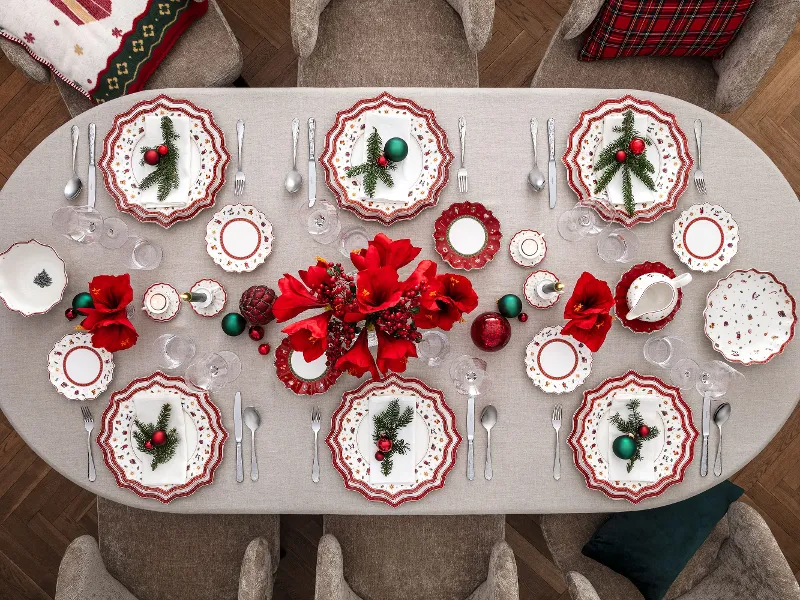 Une table de salle à manger festive dressée pour huit personnes avec des assiettes rouges et blanches Toy’s Delight de Villeroy & Boch, des couverts disposés, des fleurs rouges, des décorations de Noël et une nappe grise dans une salle à manger confortable avec des sièges gris.