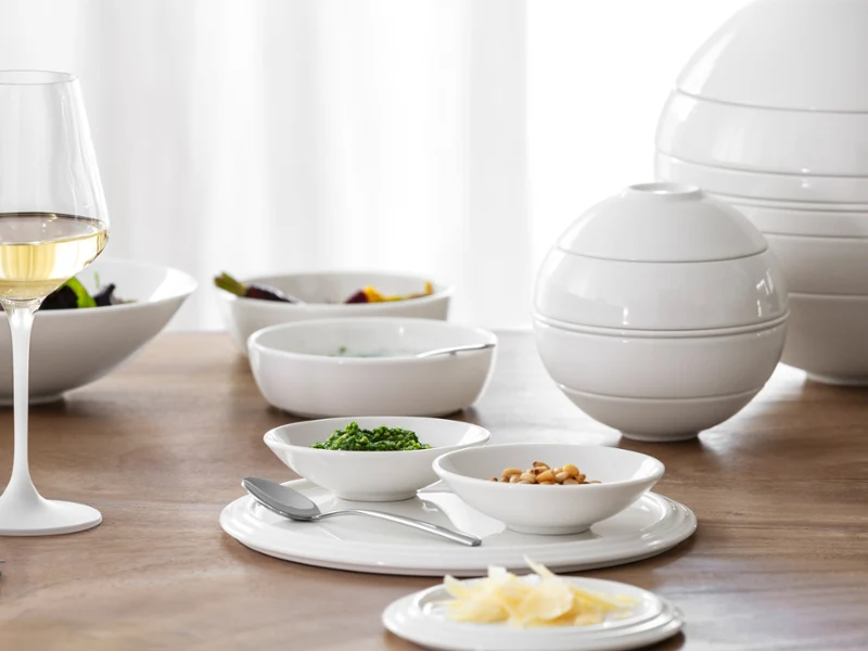 Un verre à vin blanc et de la vaisselle en céramique blanche avec de la nourriture sont disposés sur une table en bois avec une douce lumière naturelle.