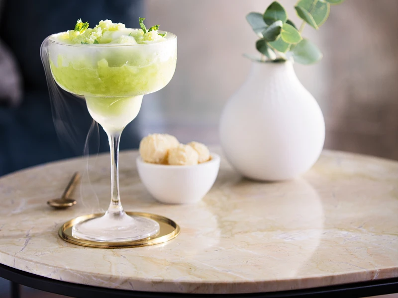 A chilled cocktail, garnished with lime twist, served in a Purismo cocktail glass on a marble table, accompanied by a small serving bowl of snacks and a white vase with green plants.