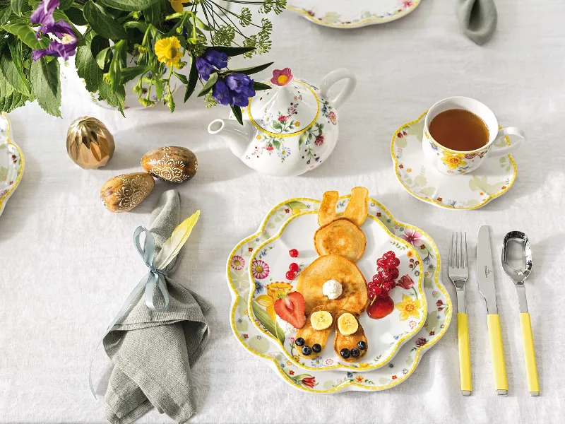 Elegante tafeldecoratie voor een feestelijke brunch met een Spring Awakening theepot met bloemenpatroon, gedessineerd Spring Awakening servies en een decoratieve haasvormige pannenkoek op een eetbord.