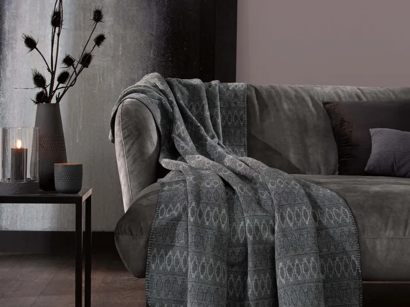 Eine graue Wohndecke auf einer grauen Couch in einem Wohnzimmer. Daneben steht ein kleiner Tisch mit einer Pflanze drauf.