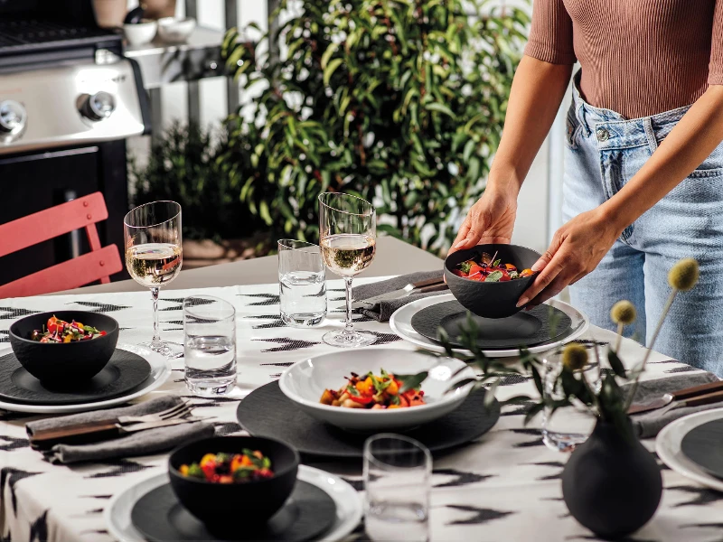 Een vrouw dekt een tafel met NewMoon en Manufacture borden en kommen voor een barbecue-avond buiten.