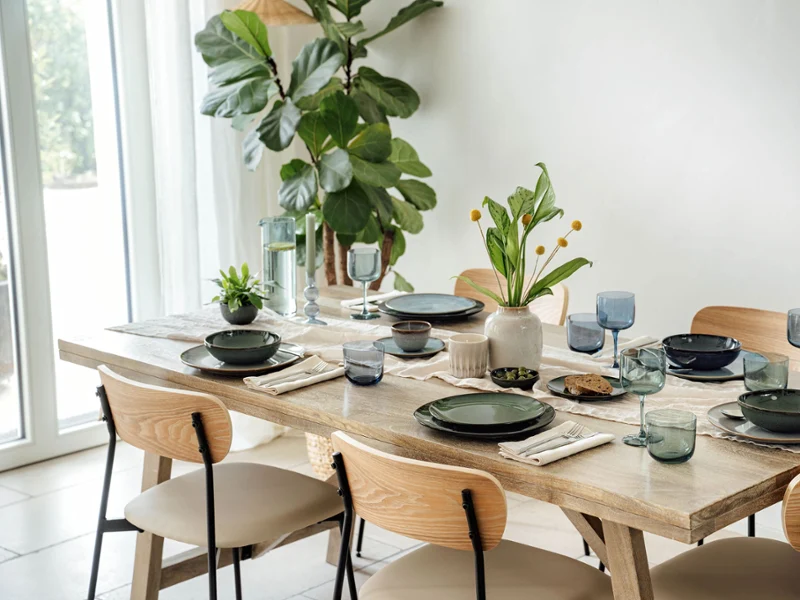 Dining table set with various items from the Lave collection by Villeroy & Boch The dining table set with various items from the Lave collection by Villeroy & Boch in green and grey, accentuated by blue glasses and napkins. A vase of yellow flowers and a small potted plant provide decoration, while a large plant stands by the window.