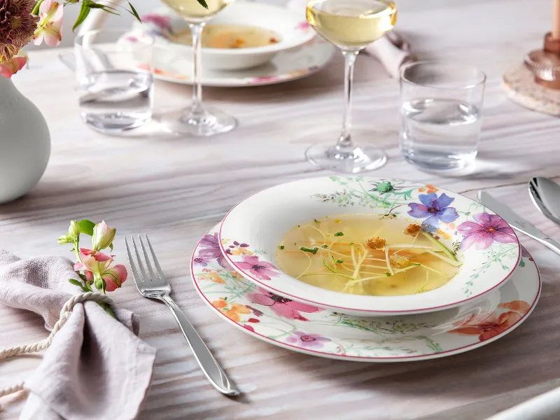 Un set da tavola con piatti dal motivo floreale "Mariefleur" di Villeroy & Boch, una ciotola per la zuppa, posate (forchetta, cucchiaio, coltello), un tovagliolo, due bicchieri da vino, due bicchieri d'acqua e un vaso con fiori.