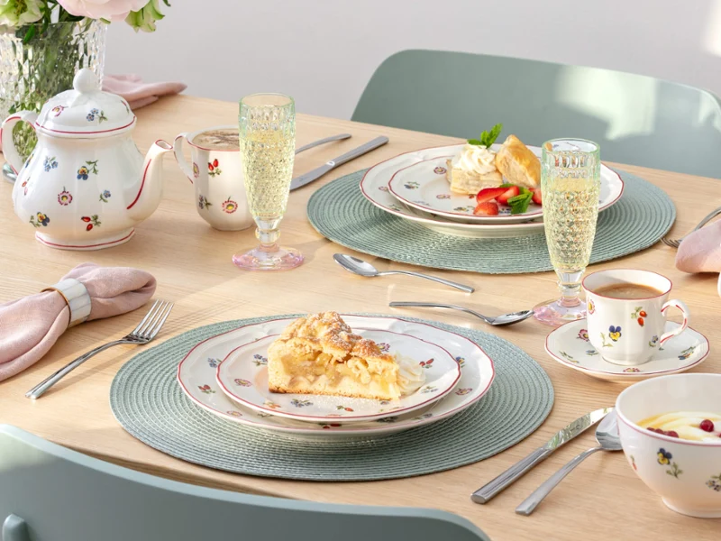 Une table dressée pour deux personnes avec des assiettes à dessert, des tasses à thé, une théière, des flûtes à champagne et de la porcelaine à motifs floraux de Petite Fleur. 