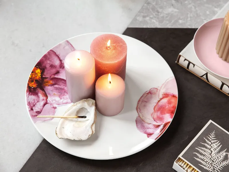 Piatto decorativo Rose Garden con candele accese accanto a una conchiglia e a un libro con motivo a foglia d'oro.