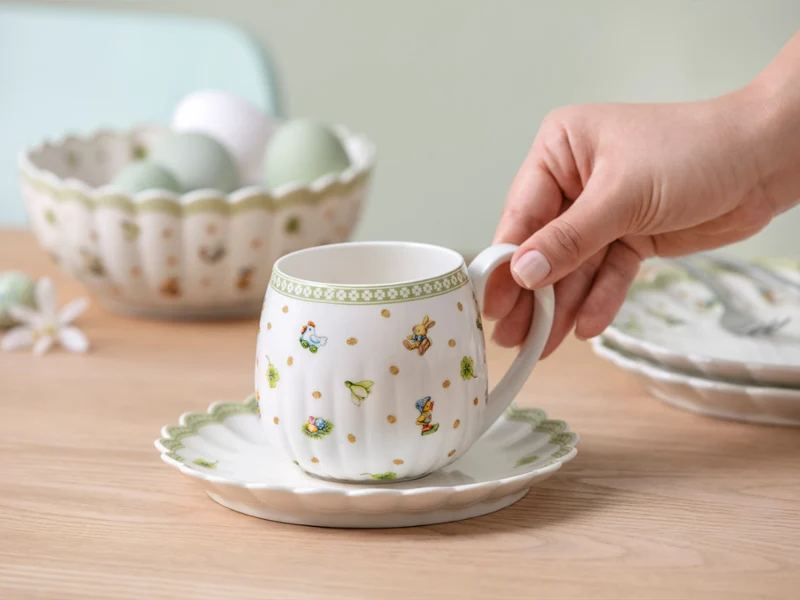 A hand holds a Easter Delight teacup on a saucer, with a bowl of eggs and plates in the background on a wooden table.