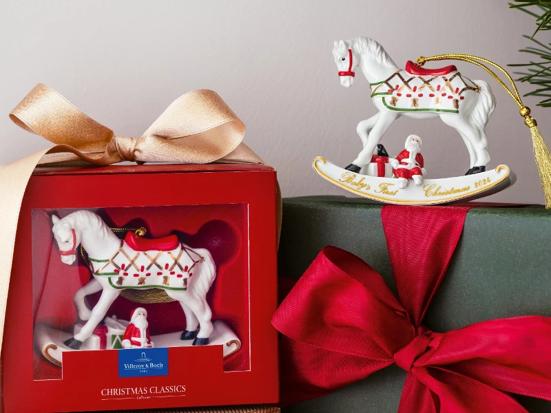 A festive presentation of two gift boxes: one with a Villeroy and Boch ornament with a rocking horse motif and another wrapped in green paper with a red bow. The Villeroy and Boch rocking horse ornament adorned with Christmas decorations can be seen above.