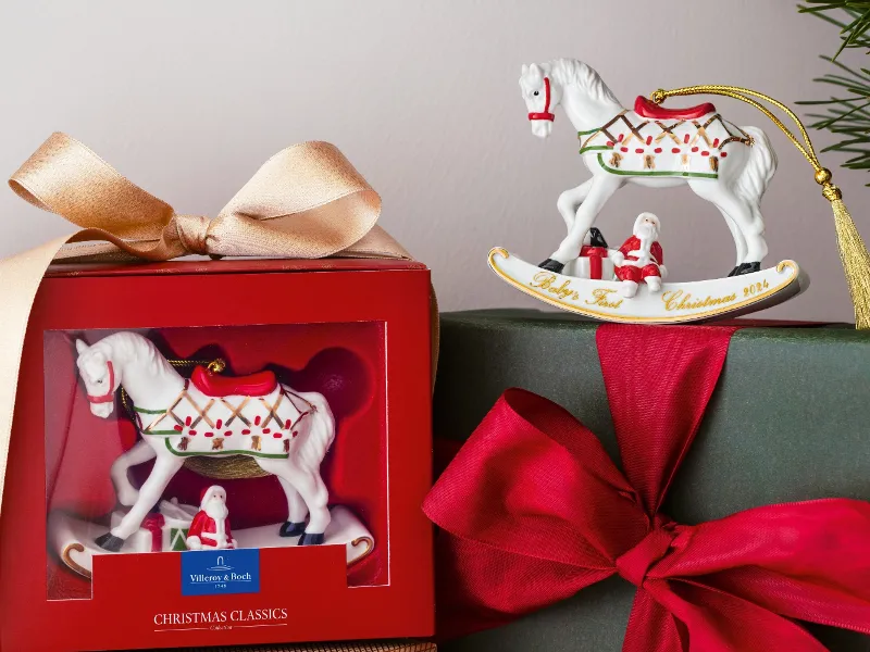 A festive presentation of two gift boxes: one with a Villeroy & Boch ornament with a rocking horse motif and another wrapped in green paper with a red bow. The rocking horse ornament from Villeroy & Boch, decorated with Christmas ornaments, can be seen above.