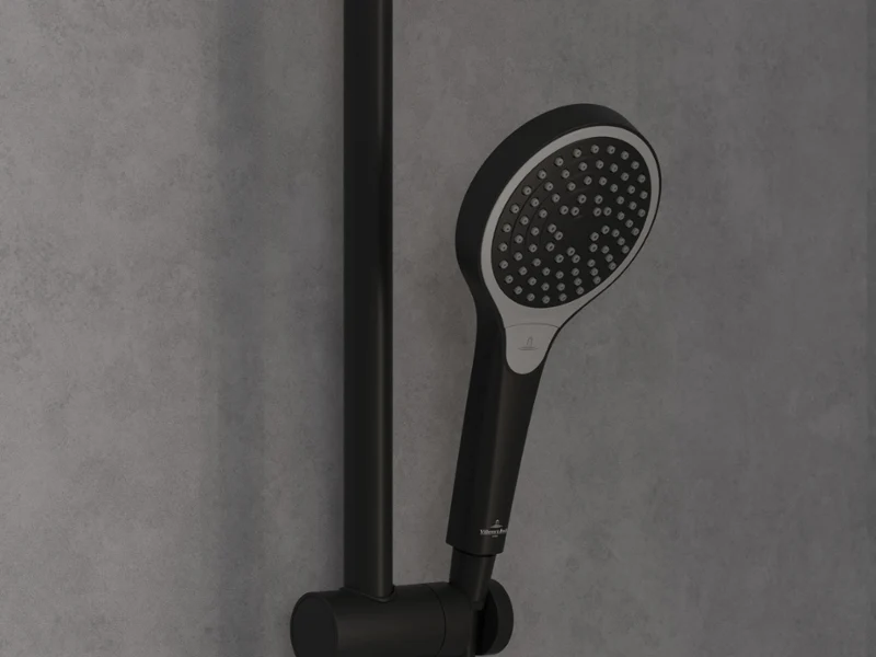 Une robinetterie de douche Verve Shower en or luxueux Une douche moderne avec une pomme de douche à effet pluie dorée et une douchette à main, montée sur un mur carrelé gris, à côté d’une tablette avec des articles de toilette et une éponge.